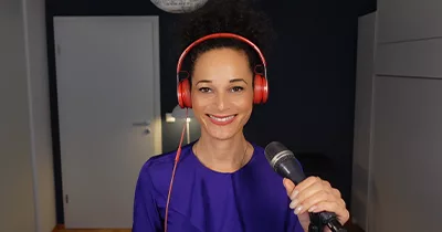 Stimmcoach Ariane Roth in lila Bluse mit roten Kopfhörern steht lächelnd am Mikrofon.
