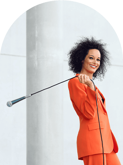 Stimmcoach Ariane Roth in orangenem Hosenanzug schaut lachend in de Kamera während sie über die rechte Schulter ein Mikrofon mit langem Kabel schwingt.